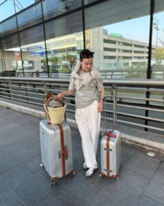 Mulher jovem e magr com duas malas de viagem. Ela usa aero look com camiseta, calça de alfaiataria e adidas samba