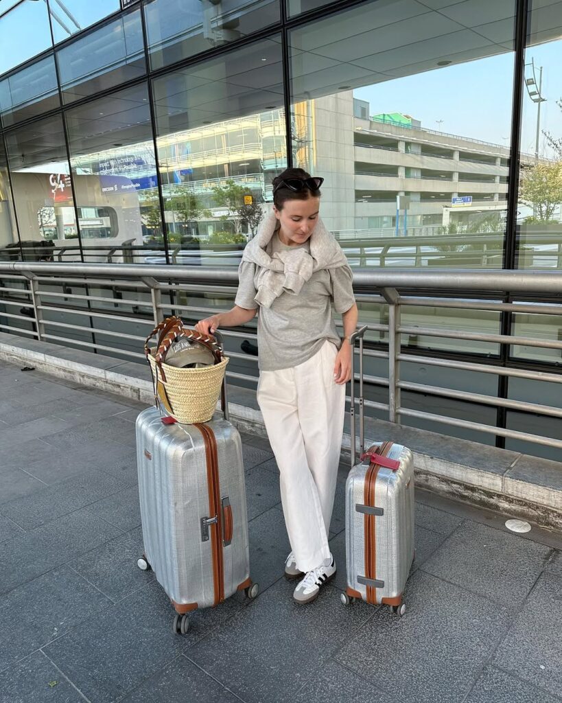 Mulher jovem e magr com duas malas de viagem. Ela usa aero look com camiseta, calça de alfaiataria e adidas samba