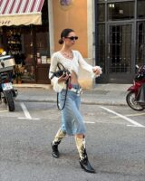 Mulher jovem e magra caminhando na rua. Ela usa blusa de manga longa cropped, jorts, cinto branco, bota western, óculos de sol e bolsa preta