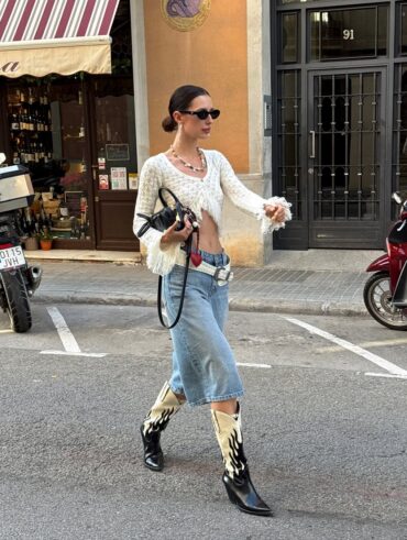 Mulher jovem e magra caminhando na rua. Ela usa blusa de manga longa cropped, jorts, cinto branco, bota western, óculos de sol e bolsa preta