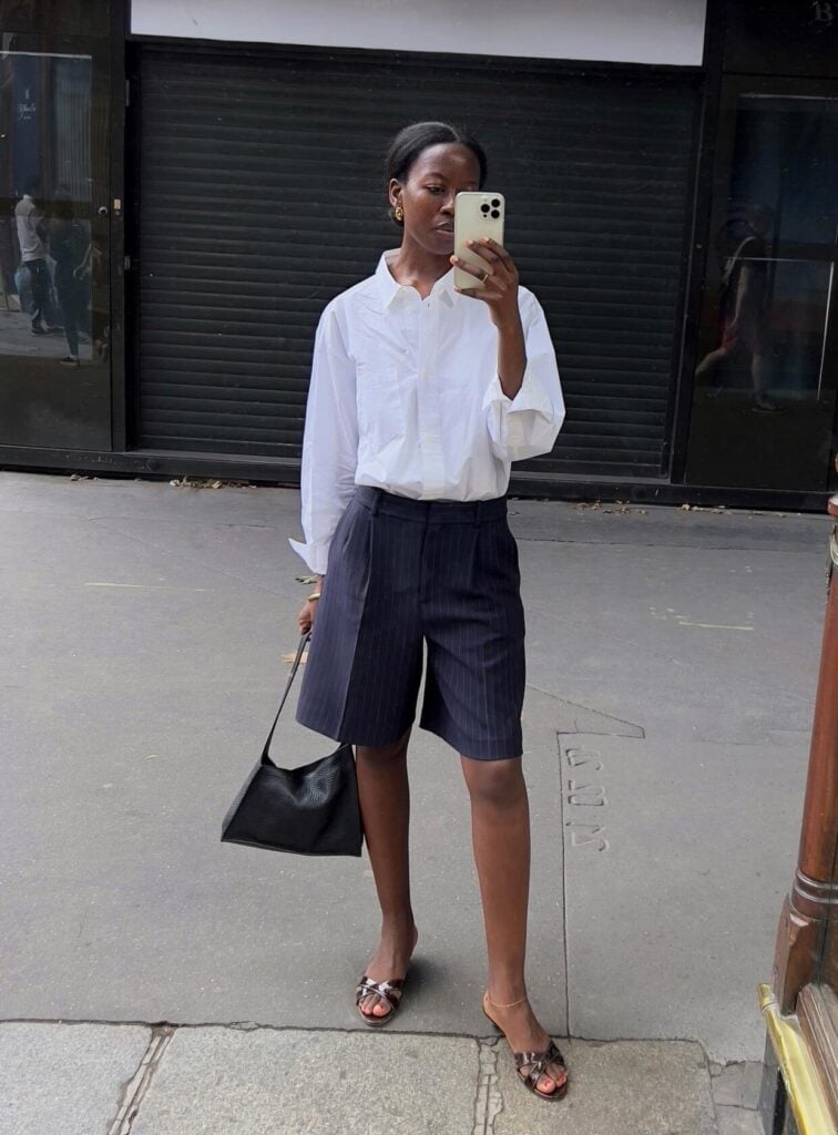 mulher jovem e magra de pele negra. Ela usa camisa branca, bermuda de alfaiataria preta e tamanco. Looks de trabalho