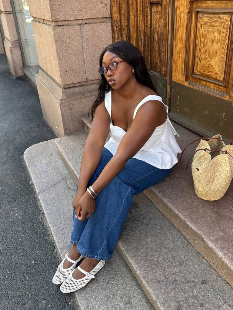 Mulher jovem de pele negra sentada em escada. Ela usa blusa branca, calça jeans e sapatilha de telinha.