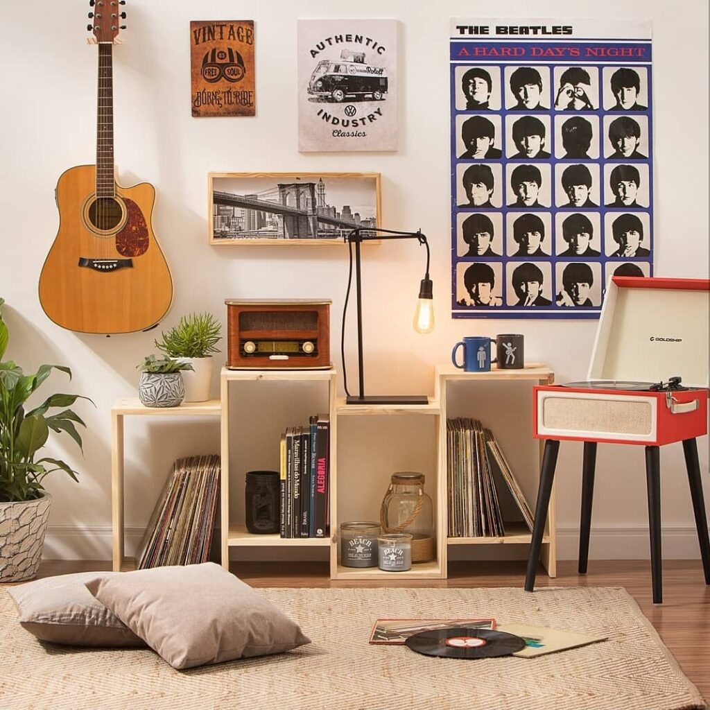 Sala de estar com vitrola vermelha e pés palito, estante com discos e rádio, poster dos Beatles e violão pendurado na parede