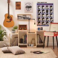 Sala de estar com vitrola vermelha e pés palito, estante com discos e rádio, poster dos Beatles e violão pendurado na parede