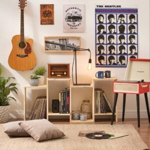 Sala de estar com vitrola vermelha e pés palito, estante com discos e rádio, poster dos Beatles e violão pendurado na parede