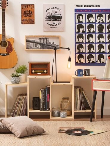 Sala de estar com vitrola vermelha e pés palito, estante com discos e rádio, poster dos Beatles e violão pendurado na parede
