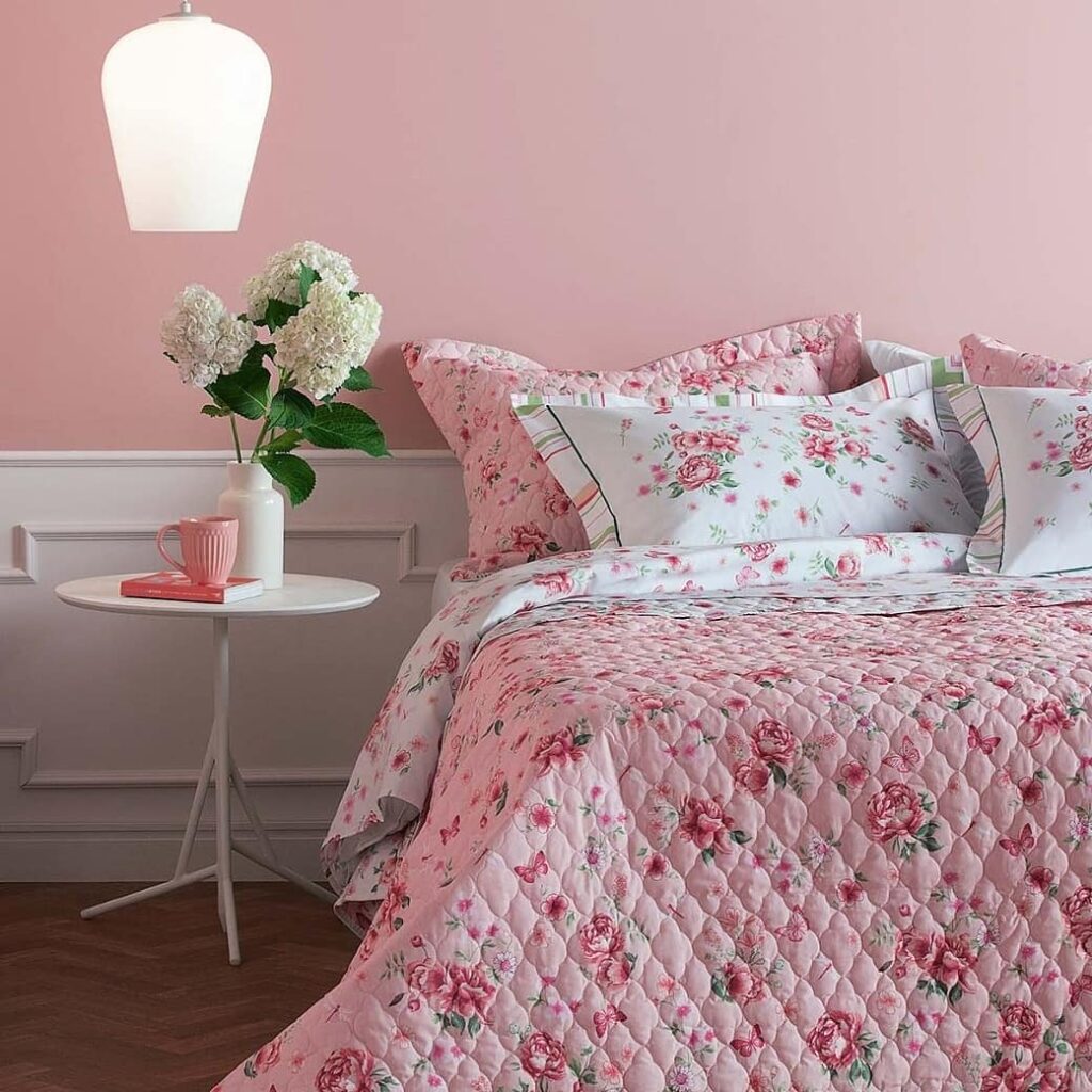 Quarto com meia parede rosa, mesa de cabeceira branca, cama posta com estampa floral