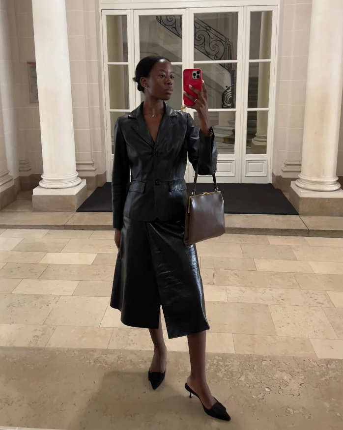 Mulher de pele preta, com blazer preto, saia preta, salto baixo e bolsa marrom