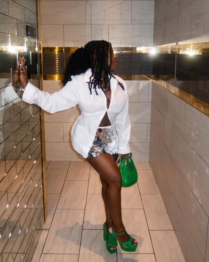 Mulher de pele preta, com camisa branca, saia de paetê prata e calçado e bolsa em verde