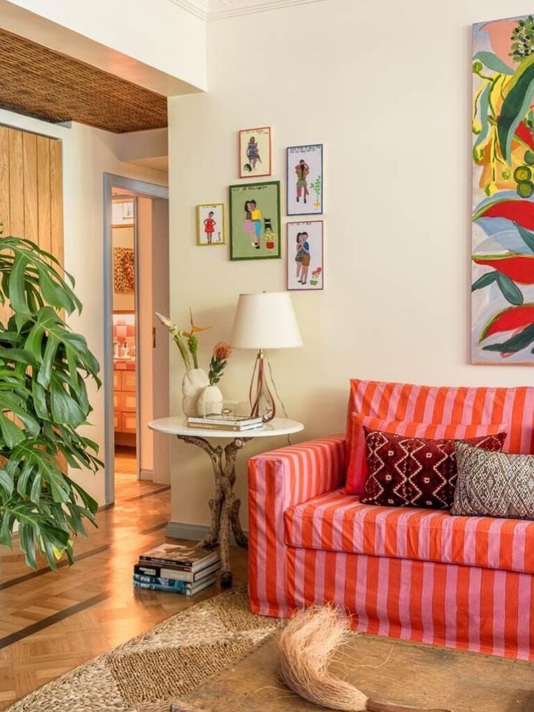 Sala de estar com sofá listrado em rosa e laranja e mesa lateral alta, redonda, com abajur e objetos decorativos