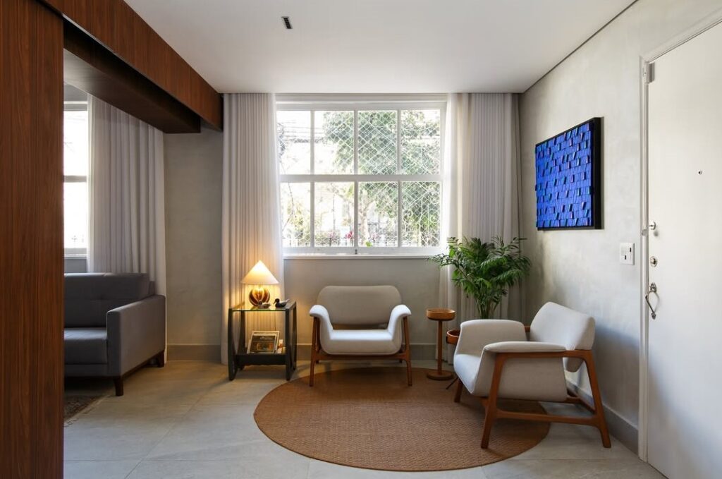 Sala de estar com poltrona estofadas bege em estrutura de madeira, tapete redondo e mesa lateral com luminária