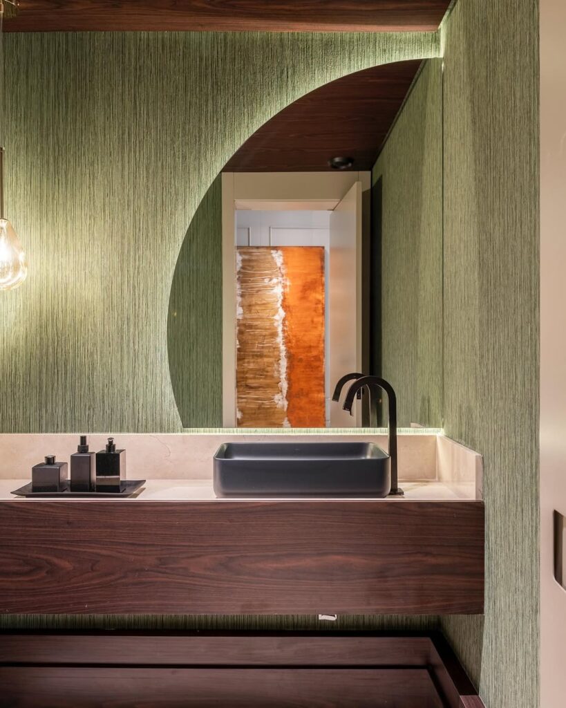 Banheiro com meio espelho circular, luz de led e papel de parede verde com textura