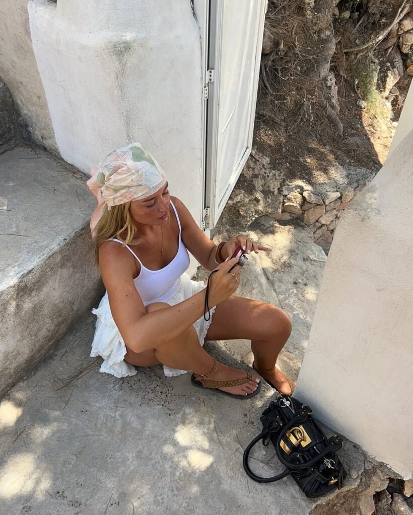 Mulher branca e magra com look boho, regata branca, lenço na cabeça, saia de renda e rasteirinha boho com pedraria e bolsa preta