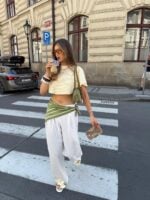 Mulher branca e magra usando calça branca, cropped listrado amarelo manteiga e branco, lenço verde na cintura, bolsa verde e sandália rasteirinha de flor