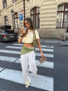 Mulher branca e magra usando calça branca, cropped listrado amarelo manteiga e branco, lenço verde na cintura, bolsa verde e sandália rasteirinha de flor