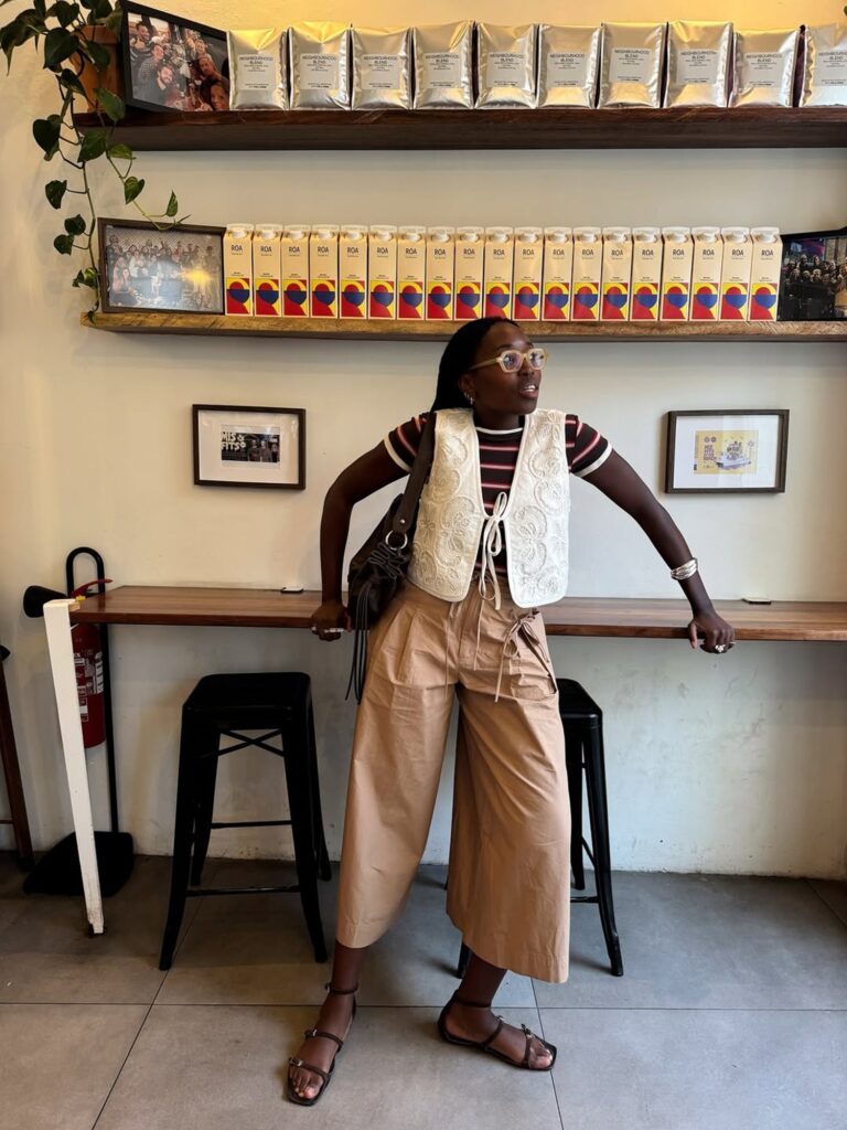 Mulher negra e magra com look de blusa listrada, calça bege, colete, bolsa e sandália rasteirinha