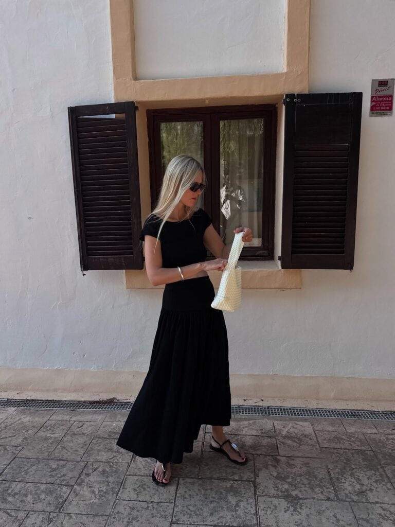 Mulher branca e magra com vestido preto longo, bolsa branca, óculos de sol e rasteirinha t-strap com metal