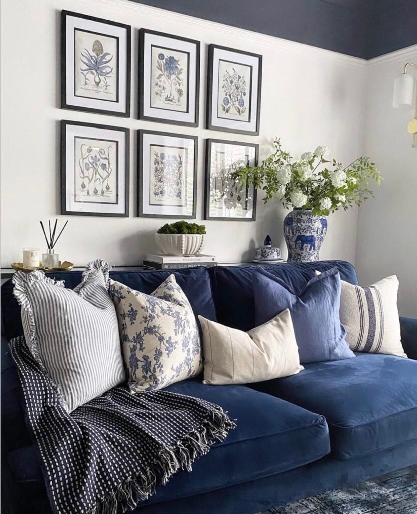 Sala com parede branca, teto escuro, sofá azul em veludo com várias almofadas e quadros com bordas pretas