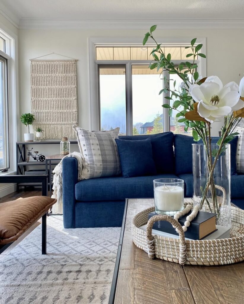 Sala com sofá azul escuro em frente à uma janela, mesa de centro de madeira com bandeja com vela, livro e vaso com flores