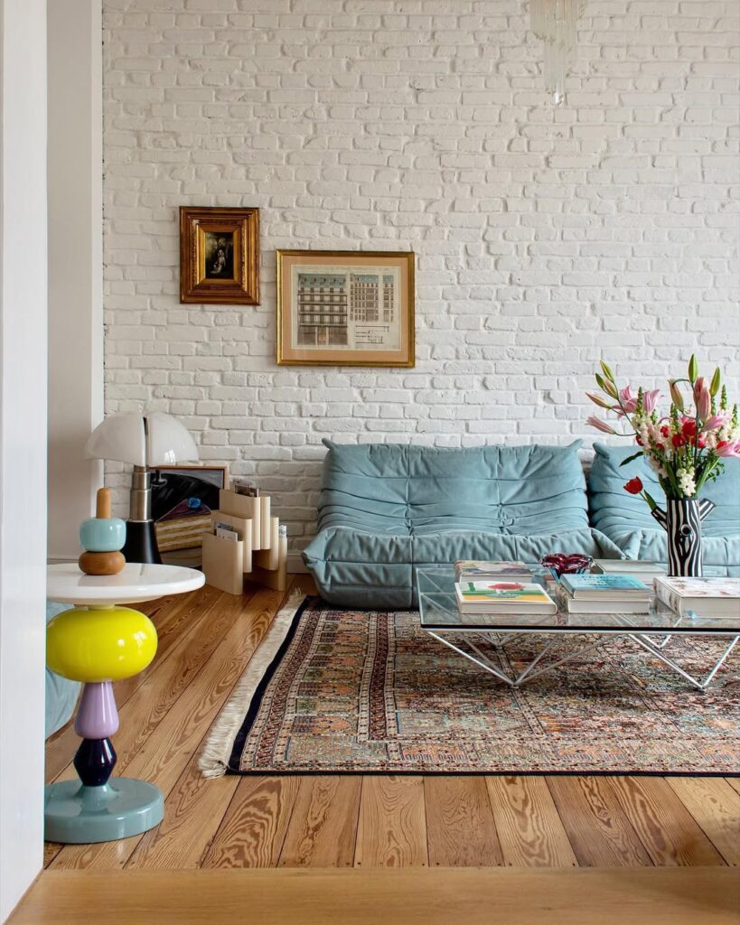 Sala de estar com pé direito alto, parede de tijolinhos brancos, sofá azul com design diferente, mesa de centro de vidro com livros e vaso de flores