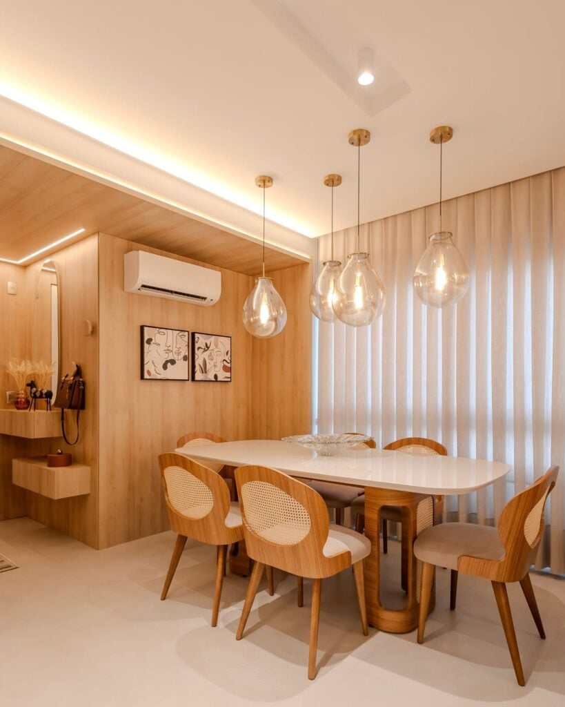 Sala de jantar com mesa branca oval com detalhes amadeirados, cadeiras com assento estofado e base em madeira, e pendentes transparentes que lembram balões