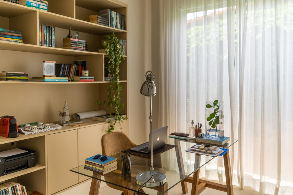 Escritório com estante na cor clara com livros e diferentes objetos decorativos, mesa com tampo de vidro e estrutura em madeira