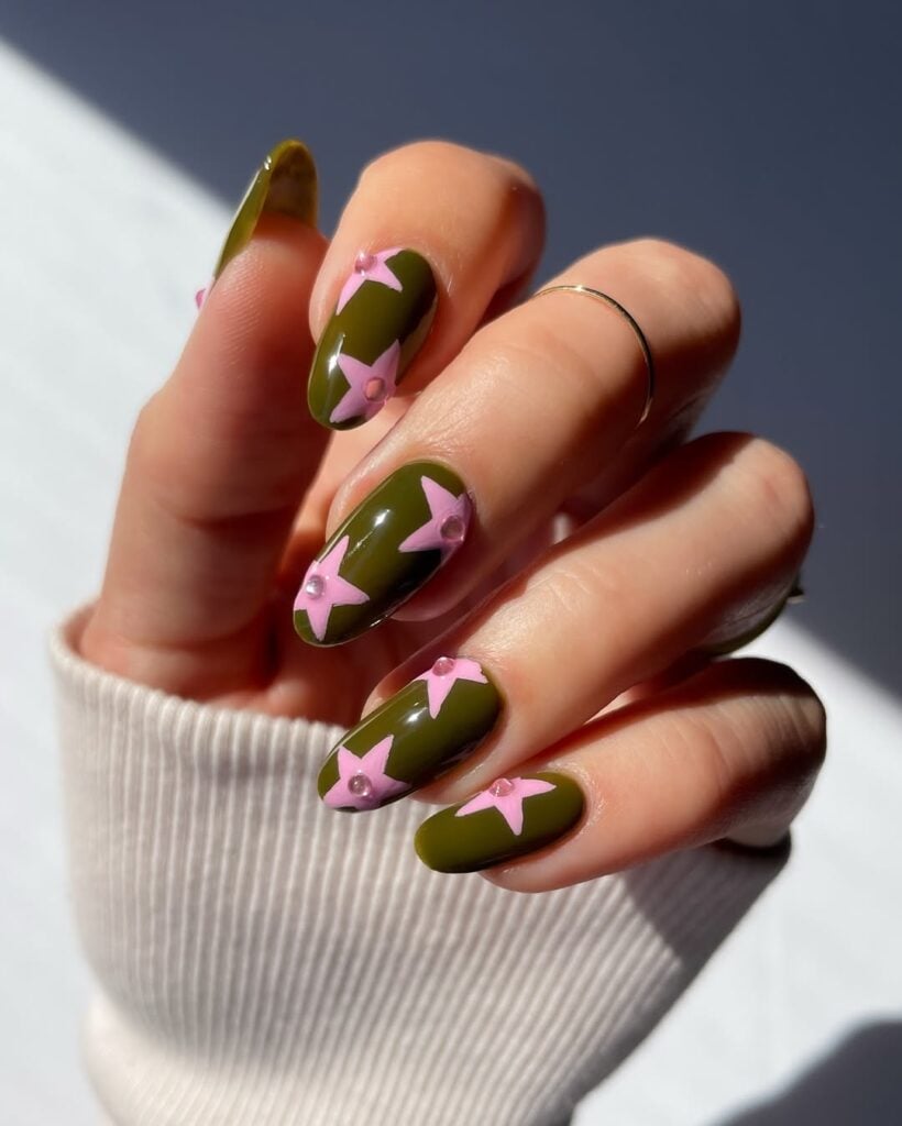 Mão de pele clara, com anel dourado e unha amendoada verde com estrela rosa 