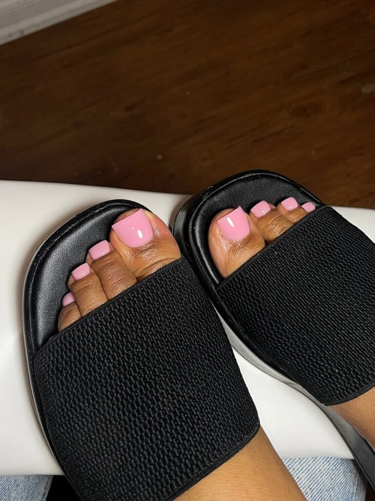 Pés de pele preta, com chinelo preto e unha rosa-claro