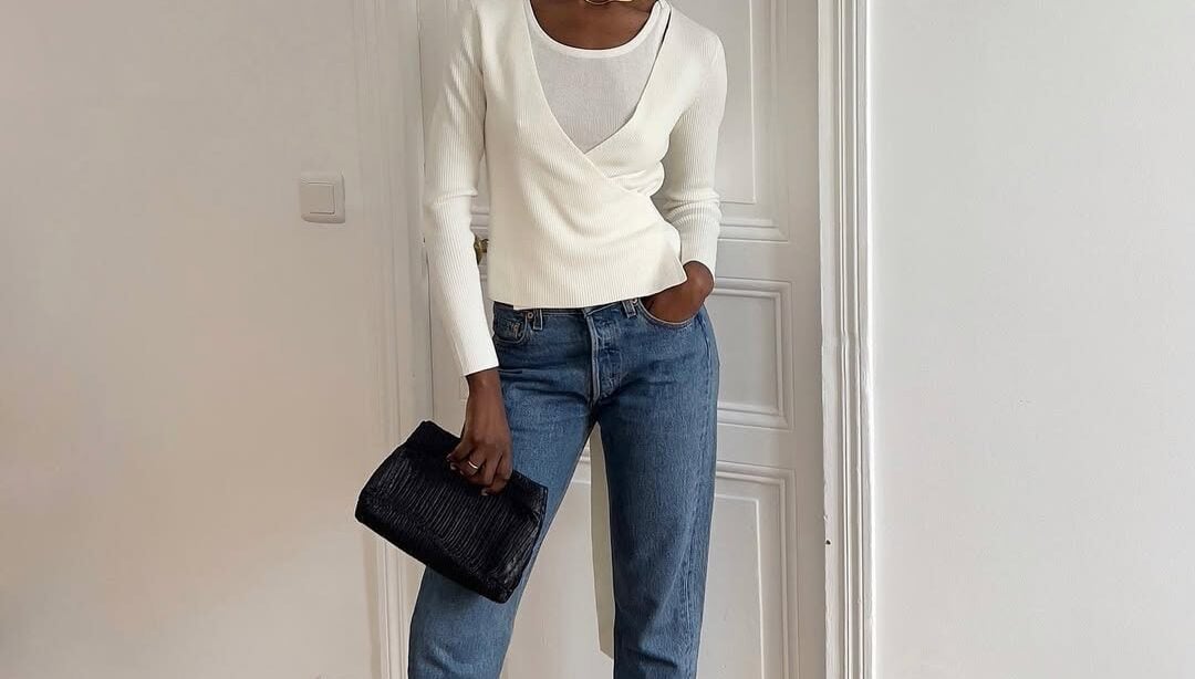 Mulher jovem e magra de pele preta. Ela usa suéter branco, calça jeans, salto alto preto e bolsa carteira preta