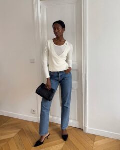 Mulher jovem e magra de pele preta. Ela usa suéter branco, calça jeans, salto alto preto e bolsa carteira preta
