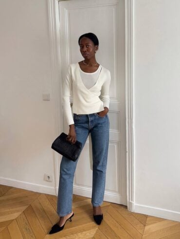 Mulher jovem e magra de pele preta. Ela usa suéter branco, calça jeans, salto alto preto e bolsa carteira preta