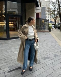Mulher jovem, magra e de pele clara. Ela vese casaco longo trench coat, blusa branca, calça jeans com sapatilha e cinto
