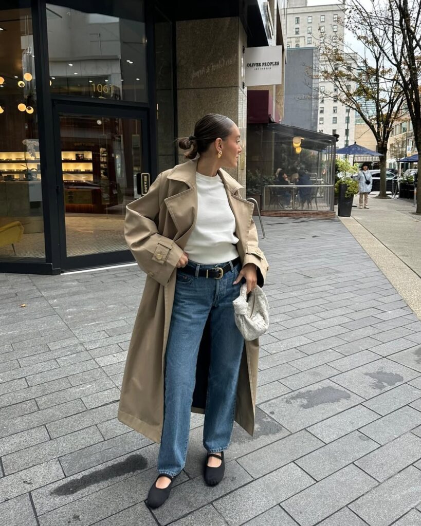 Mulher jovem, magra e de pele clara. Ela vese casaco longo trench coat, blusa branca, calça jeans com sapatilha e cinto