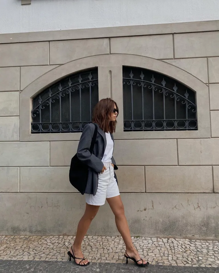 mulher jovem, magra e de pele clara com cabelo médio. Ela usa blazer, blusa, bermuda jeans branca e sandália de salto alto