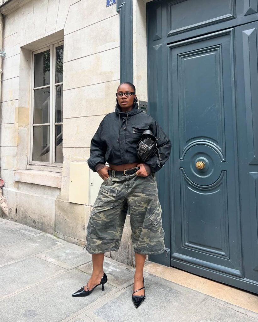 Mulher jovem de pele preta. Ela usa jaqueta, bermuda jeans longa, salto alto com bico fino e cinto