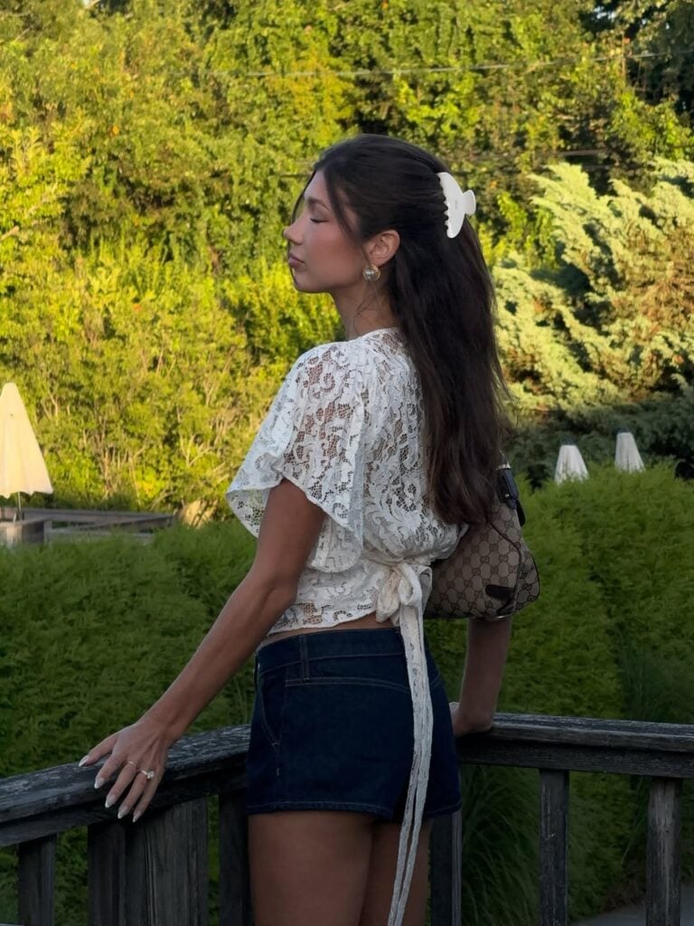 Mulher jovem, magra e de pele clara com cabelos castanhos. Ela usa blusa de renda e short jeans