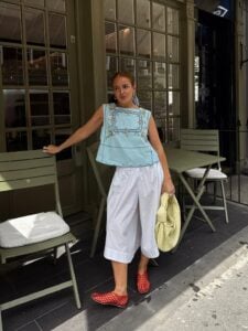 mulher jovem com blusa bata azul com detalhes bordados, calça branca, sapatilha telada vermelha e bolsa amarela