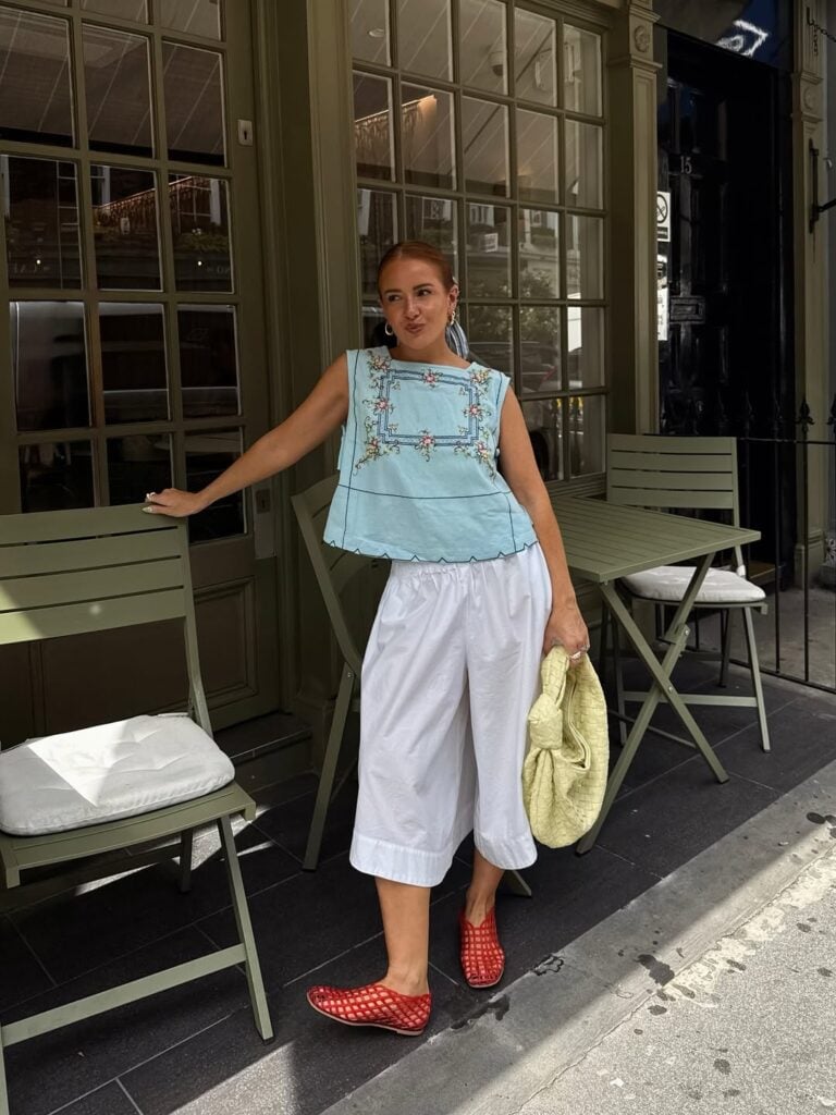 mulher jovem com blusa bata azul com detalhes bordados, calça branca, sapatilha telada vermelha e bolsa amarela
