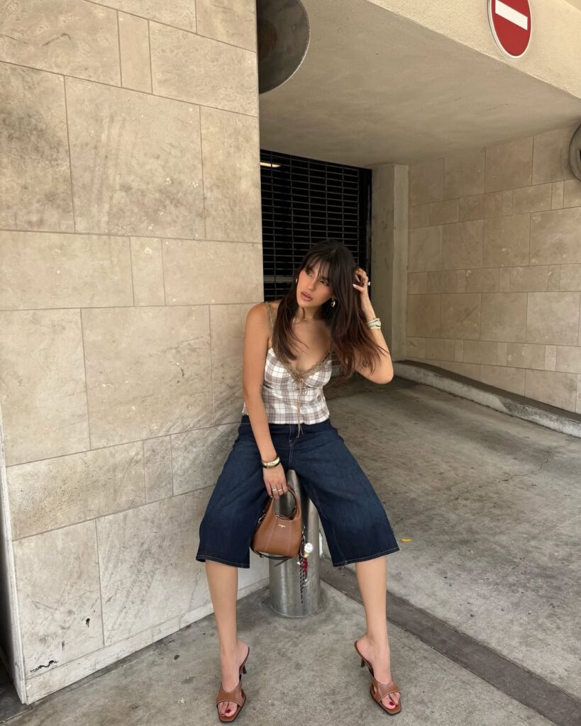 Mulher jovem e magra de pele clara. Ela usa blusa de alça, bermuda e salto alto