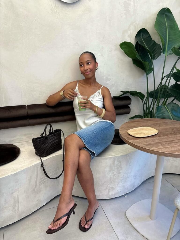Mulher jovem, magra e de pele negra. Ela usa regata acetinada e bermuda jeans com chinelo kitten heels