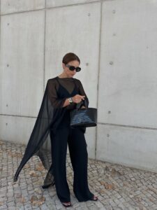 mulher jovem com pele clara e cabelo preso. Ela usa look todo preto com blusa transparente por cima