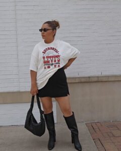 Mulher jovem, de pele clara, mid size e com cabelo castanho prezo em um coque. Ela está na frente de uma parede e usa t-shirt estampada, saia curta preta, bota de cano longo preta e bolsa preta.