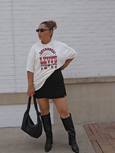 Mulher jovem, de pele clara, mid size e com cabelo castanho prezo em um coque. Ela está na frente de uma parede e usa t-shirt estampada, saia curta preta, bota de cano longo preta e bolsa preta.