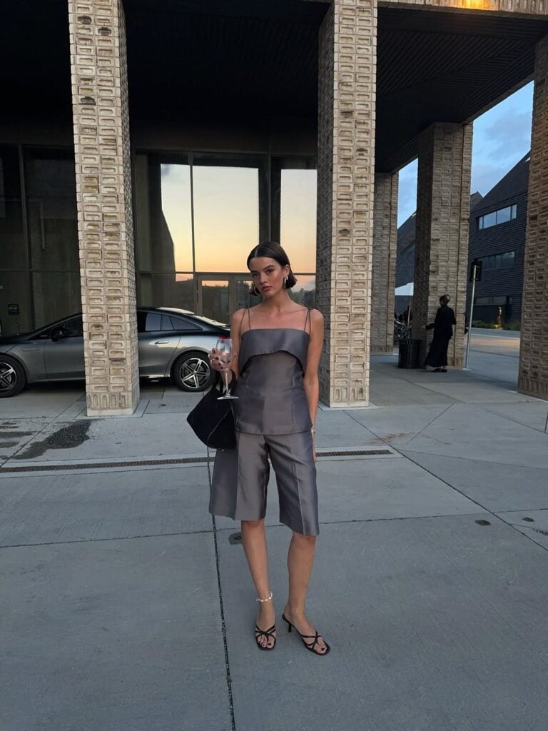 mulher jovem, magra e de pele clara. Ela usa conjunto cinza, tamanco e bolsa