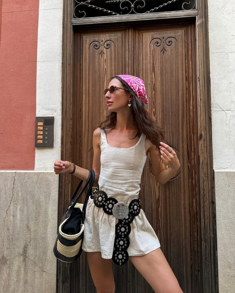 Mulher jovem, magra e de pele clara. Ela usa vestido curto com cinto boho