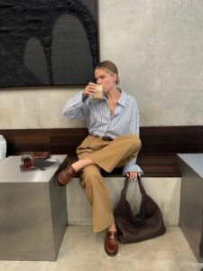 mulher jovem, magra e de pele clara com cabelos loiros. Ela usa camisa de manga longa listrada, calça caqui, mocassim marrom claro e bolsa marrom
