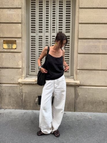 mulher jovem, magra e de pele clara. Ela usa blusa acetinada preta, calça jeans branca e sapatilha