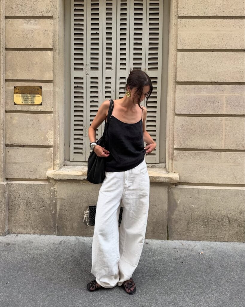 mulher jovem, magra e de pele clara. Ela usa blusa acetinada preta, calça jeans branca e sapatilha