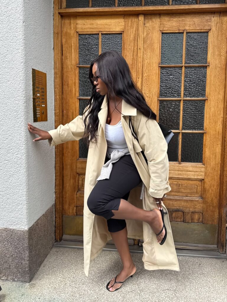 Mulher jovem de pele negra e cabelos longos. Ela usa casaco trench, regata, calça capri e chinelo kitten heels