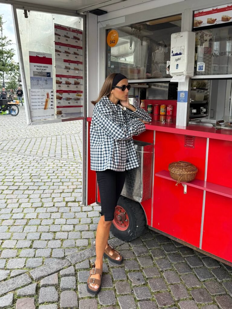 Mulher jovem, magra e de pele clara e cabelo castanho. Ela usa casaco estampado, calça capri e papete birkenstock